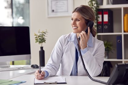 Smiling Female Doctor Or GP Wearing White Coat Sitting At Desk I Smiling Female Doctor Or GP Wearing White Coat Sitting At Desk I